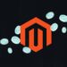 Rotes Magento-Logo in Hexagon-Form schwebt zentriert auf schwarzem Hintergrund mit dezenten Linienmustern; mehrere türkisfarbene, halbtransparente Münzen oder Scheiben fliegen diagonal über das Bild.